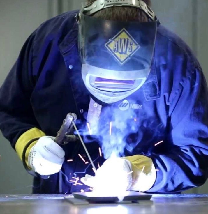 A welder in protective gear works on metal, creating sparks and bright blue light.
